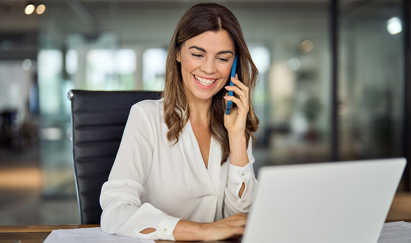 Uśmiechnięta kobieta rozmawiająca przez telefon i pracująca na laptopie w nowoczesnym biurze.