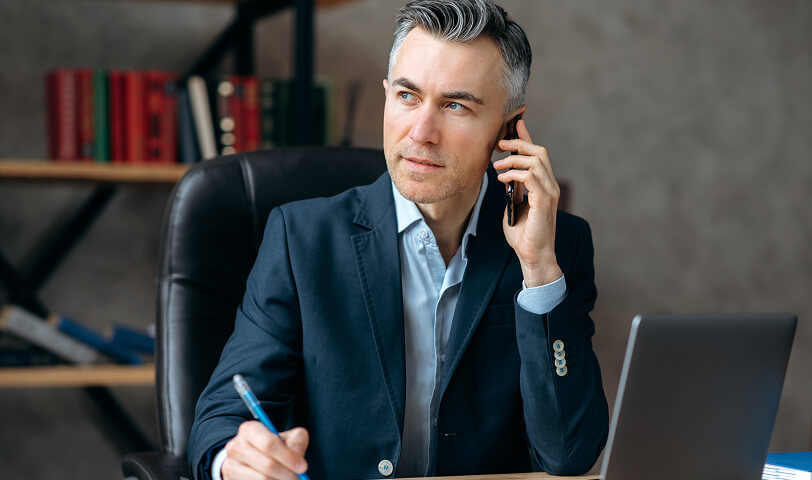 Mężczyzna w garniturze rozmawia przez telefon i robi notatki przy biurku z laptopem.