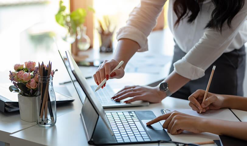 Dwie osoby siedzące przy biurku współpracują nad projektem, jedna pisze na tablecie, a druga korzysta z laptopa.